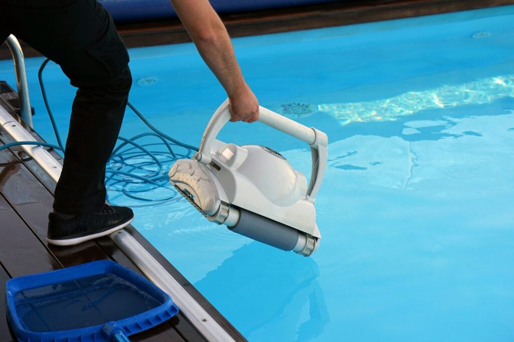 Eine Person setzt einen weißen Poolroboter vom Beckenrand ins Wasser, daneben ein Kescher