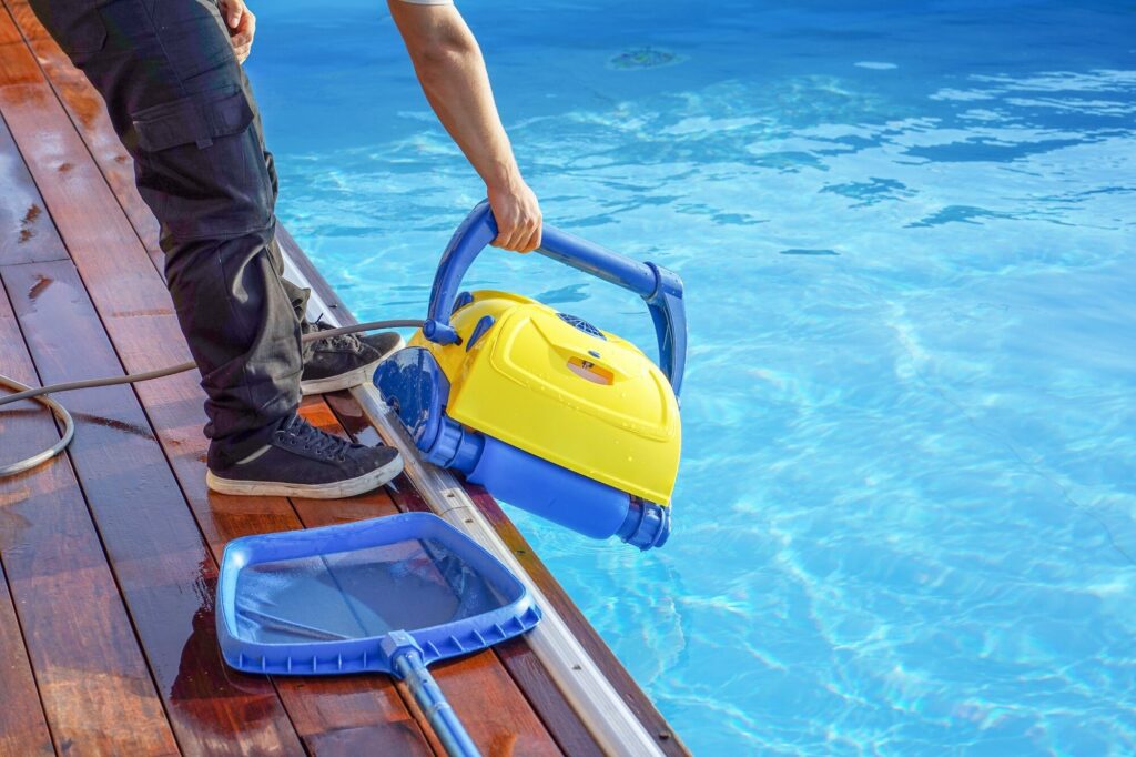 Gelber Poolroboter wird am Tragegriff aus dem Becken gehoben, daneben ein Laubkescher
